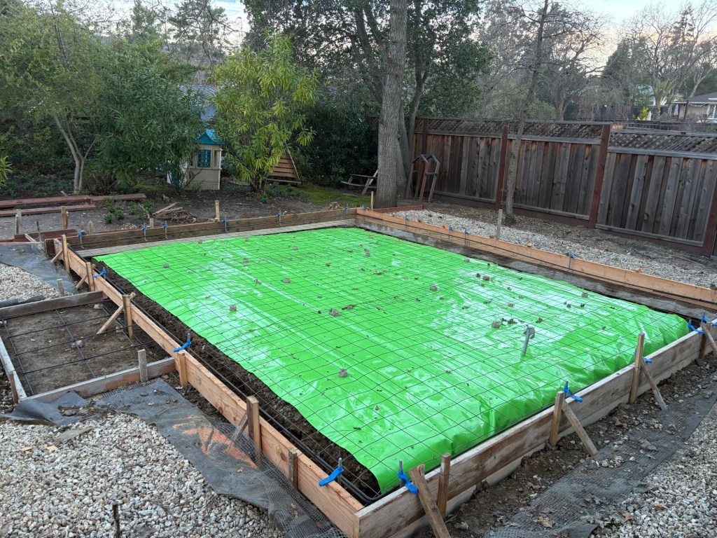 Backyard foundation forms with green vapor barrier and rebar grid laid inside, ready for the concrete pour