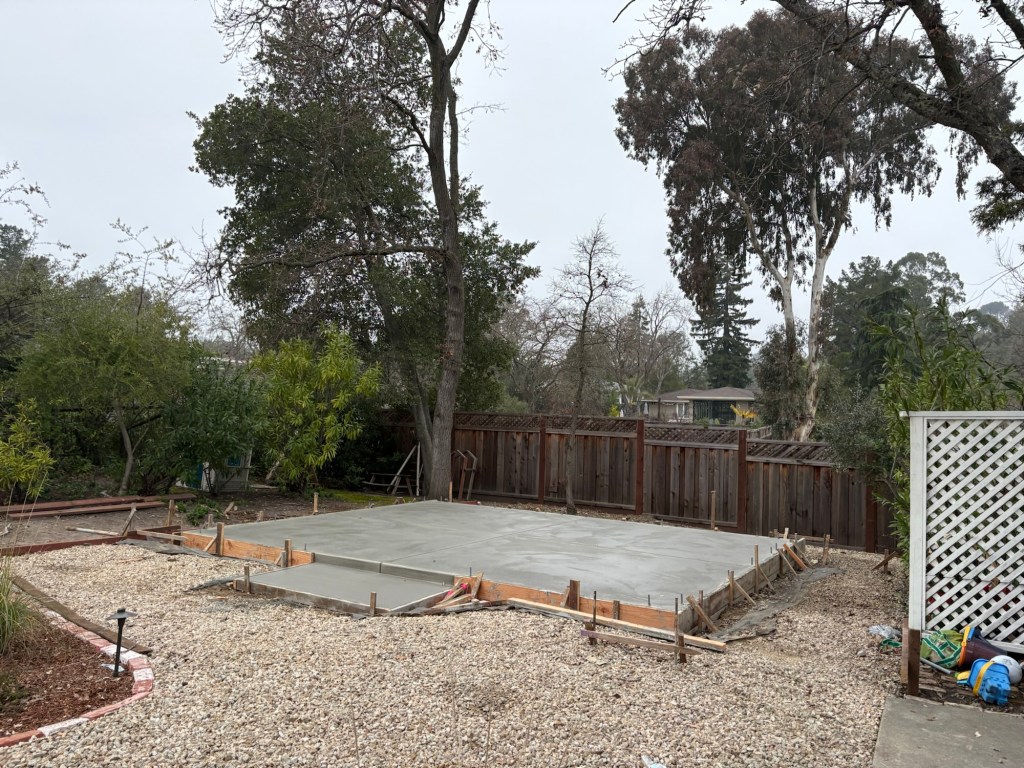 Freshly poured concrete slab still set in wooden forms in the backyard, surrounded by gravel, a wooden fence, and trees