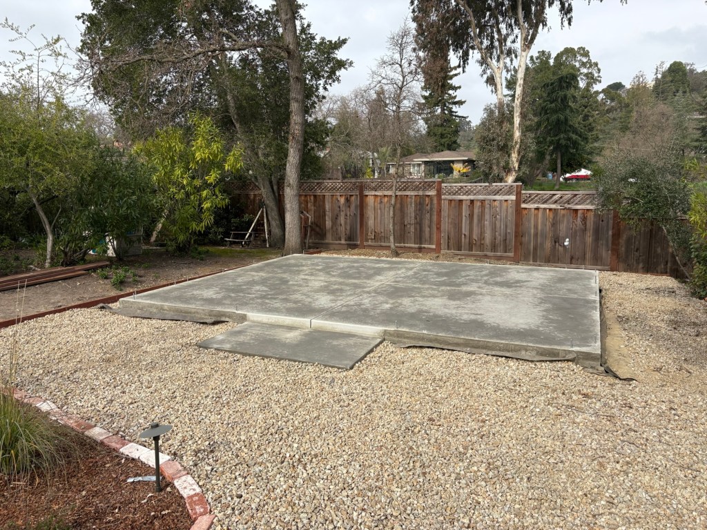 Finished concrete slab with forms removed in the backyard, surrounded by gravel, a wooden fence, and trees