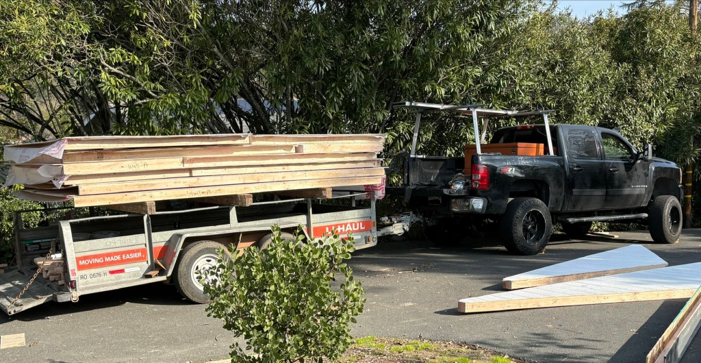 Pickup truck and trailer loaded with stacked lumber and prebuilt Tuff Shed wall sections parked in the driveway