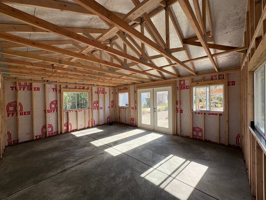 Interior of the newly installed Tuff Shed showing exposed wooden roof trusses and wall framing, Tuff Shed branded house wrap, concrete floor, and sunlight streaming through the French doors and windows
