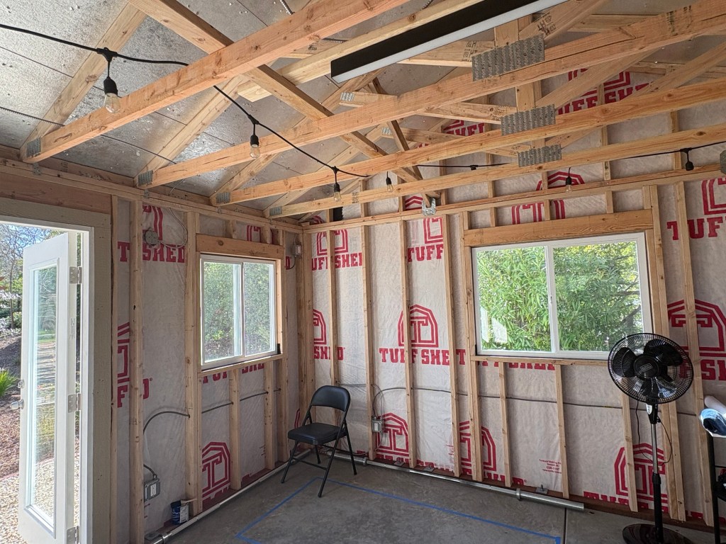 Interior of the gym shed during electrical rough-in, showing exposed wooden wall framing and ceiling trusses, metal conduit wiring along the studs, hanging string lights, and Tuff Shed house wrap on the walls