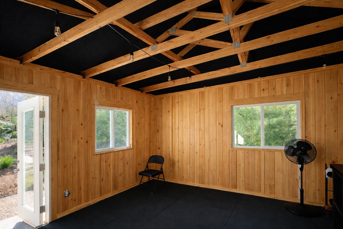 AI-generated design render of the shed gym: wood plank walls, black ceiling with open rafters, string lights, and a folding chair