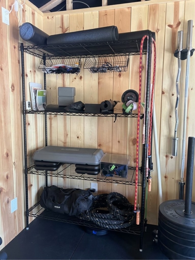 Black wire storage rack in the gym shed holding yoga mats, resistance bands, jump ropes, a step platform, a gym bag, and battle ropes.