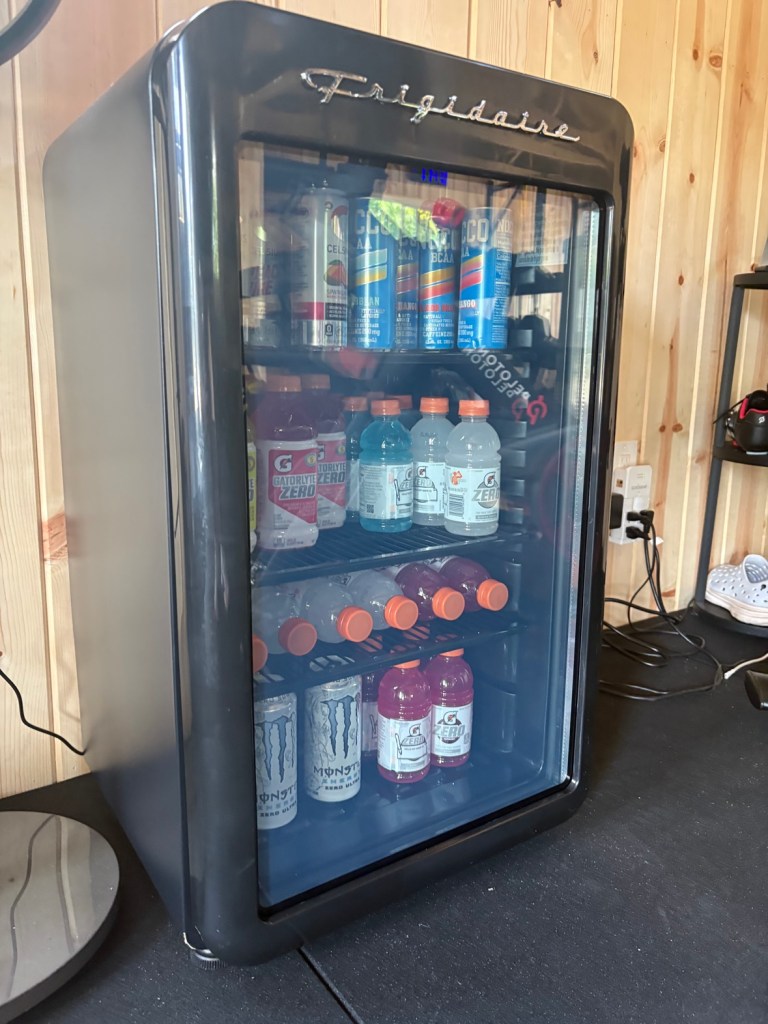 Black Frigidaire glass-door mini fridge in the gym stocked with energy drinks and sports drinks.