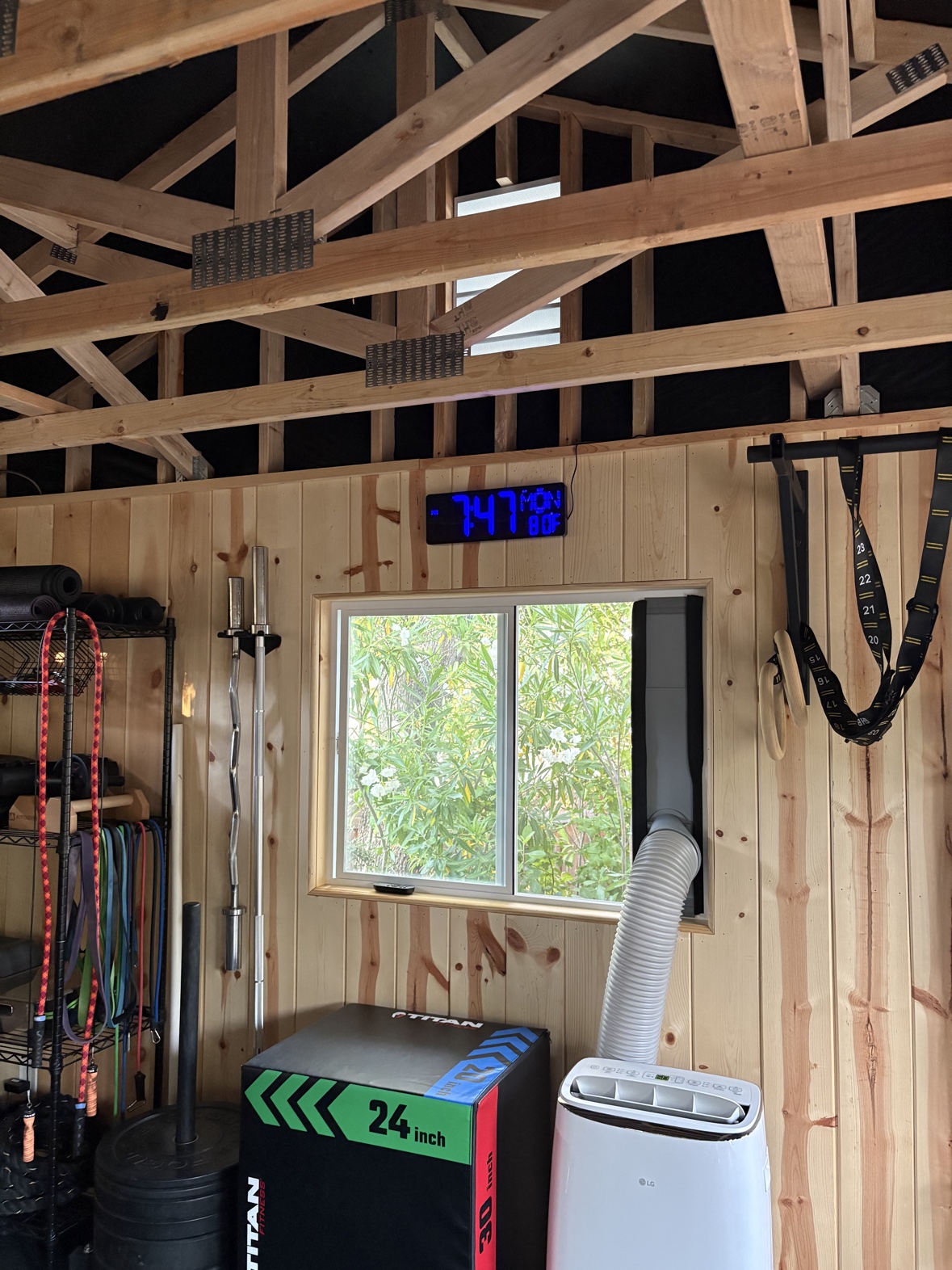 Gym shed wall featuring a window, a Titan 24-inch plyo box, an LG portable air conditioner, a wall clock, two barbells mounted on a wall hanger, gymnastic rings and TRX straps, and resistance bands.