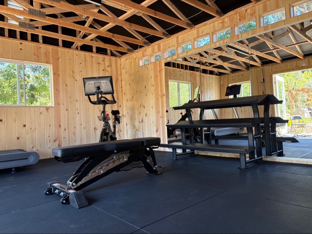 Empty black three-tier dumbbell rack beside the adjustable workout bench in the home gym, with the Peloton bike behind.