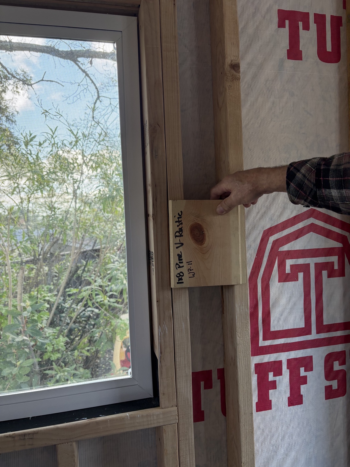 Carpenter holding a 1x8 pine V-rustic plank sample against the shed’s wall studs next to a window